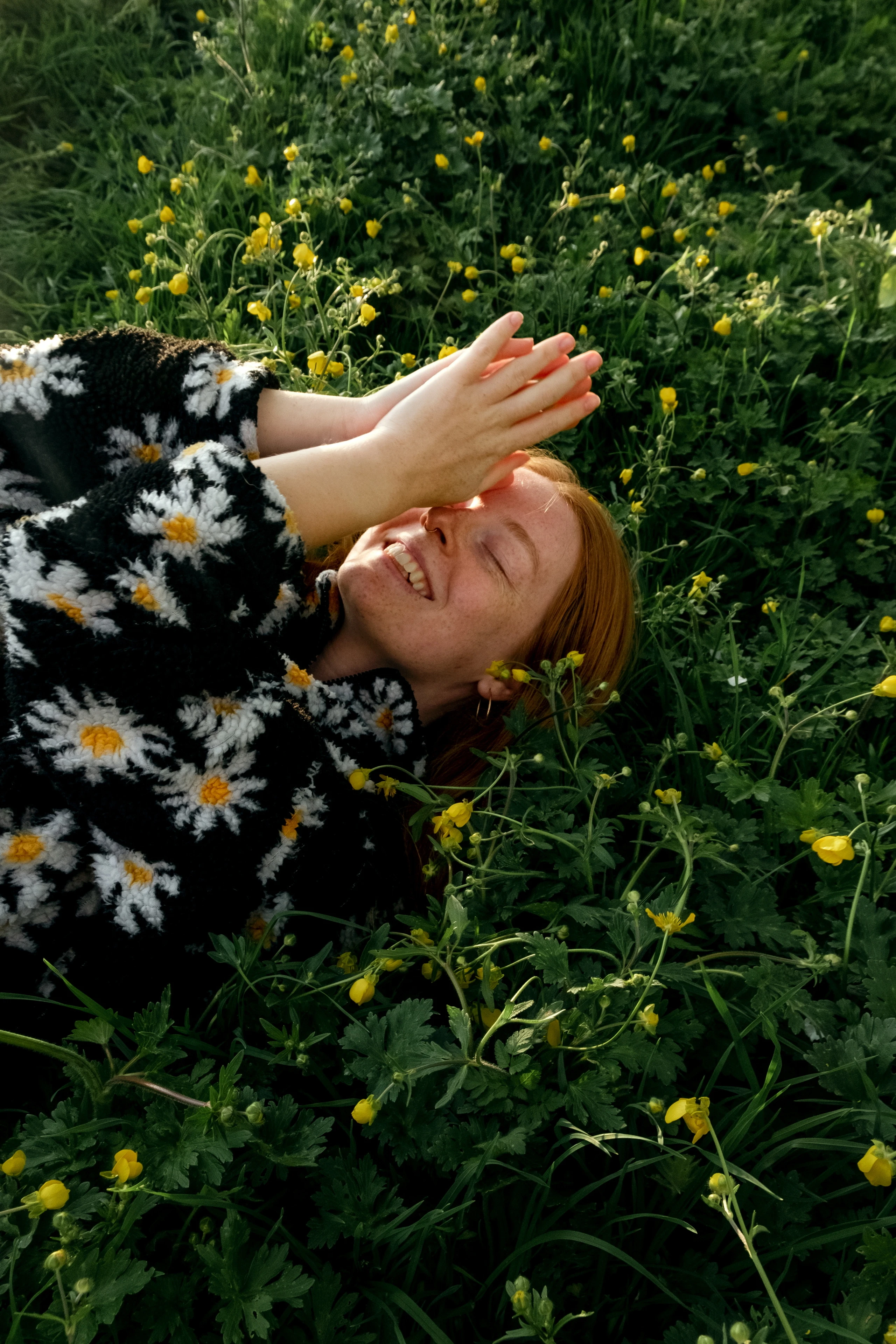 Frau mit Gänseblümchenpullover liegt in Blumenwiese und lächelt