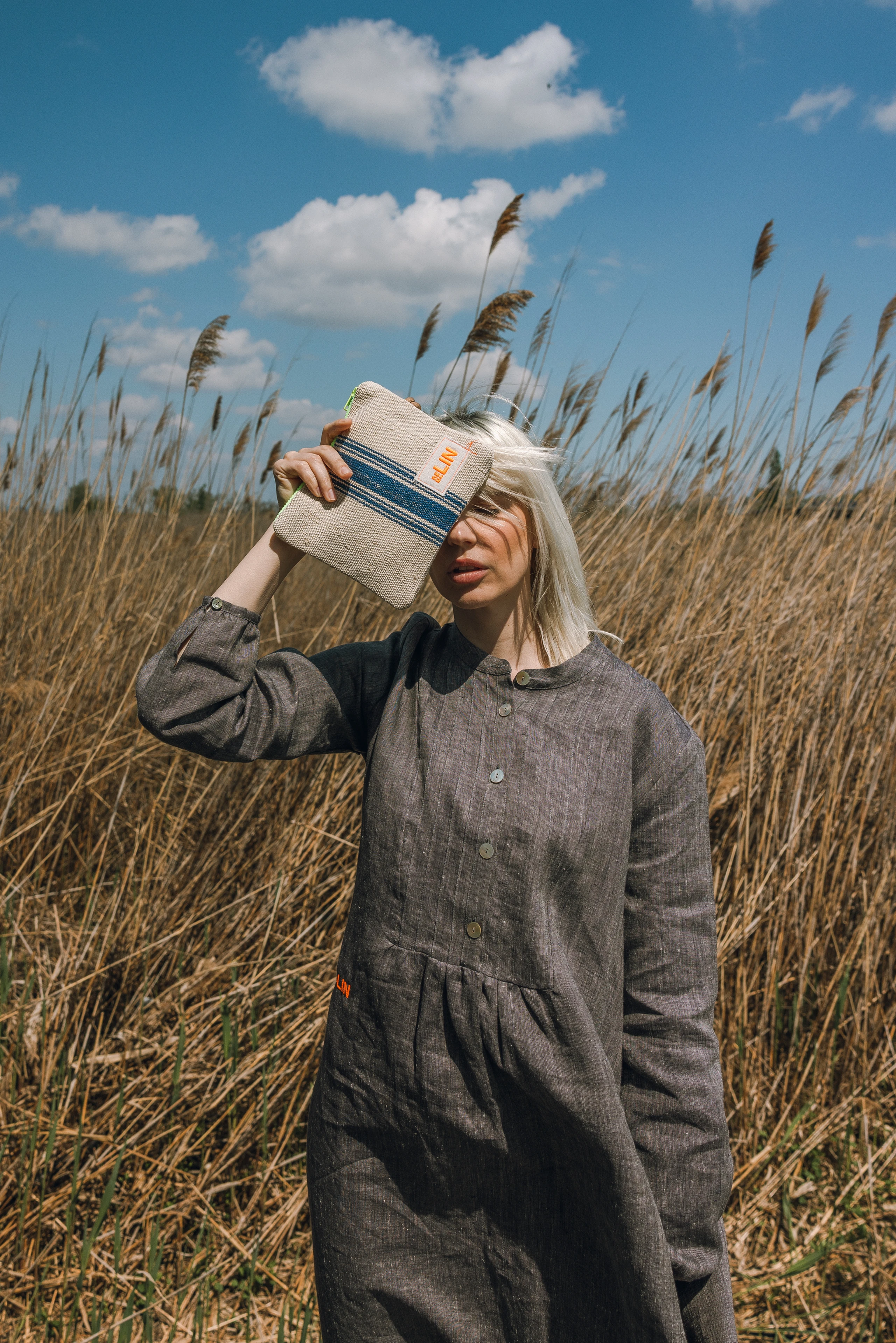Model steht in einem Feld und hält sich eine Leinentasche vors Gesicht