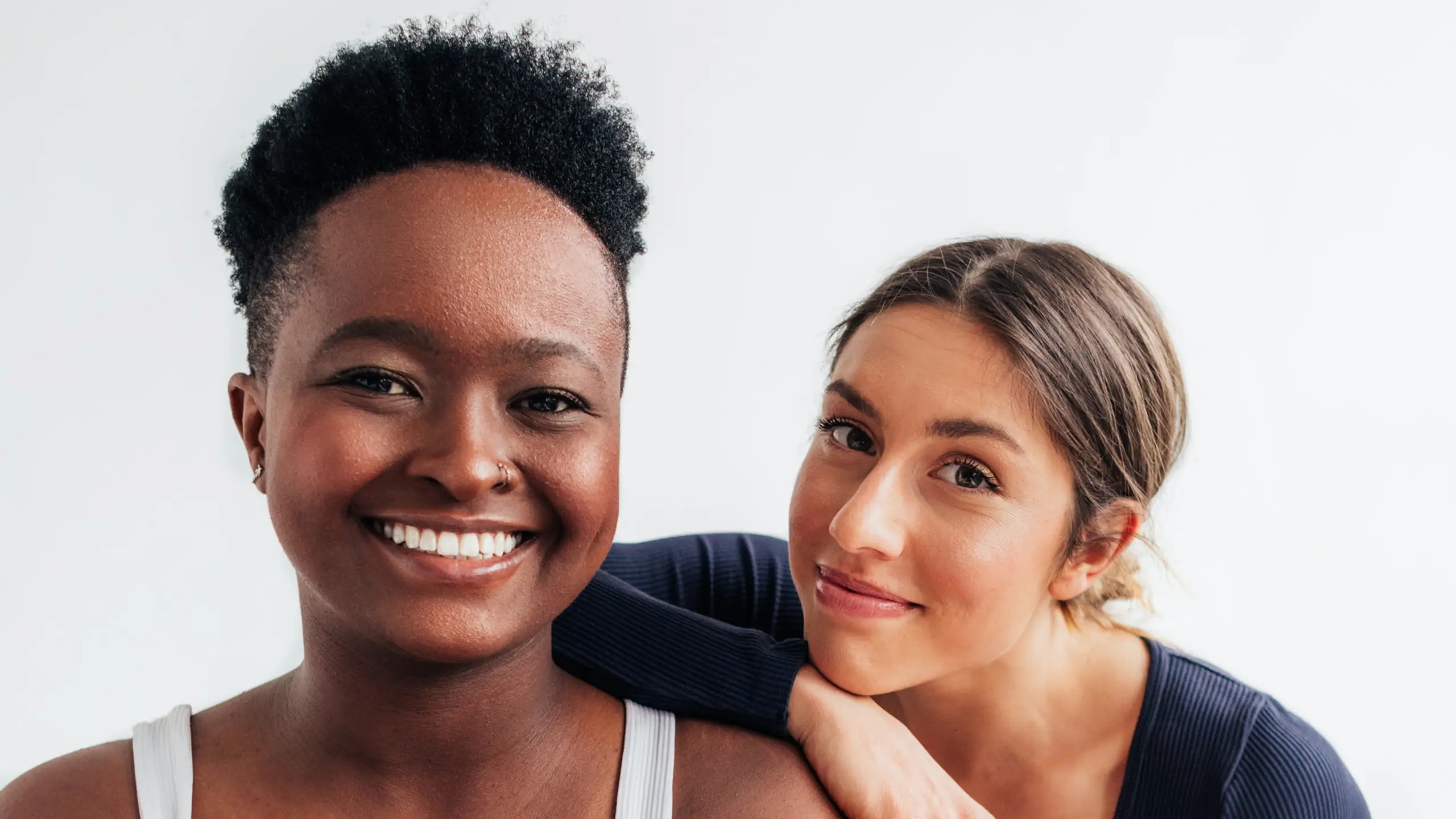 Zwei lächelnde Frauen, eine ist Schwarz und hat kurze haare, die andere ist weiß und hat längere Haare und einen Zopf
