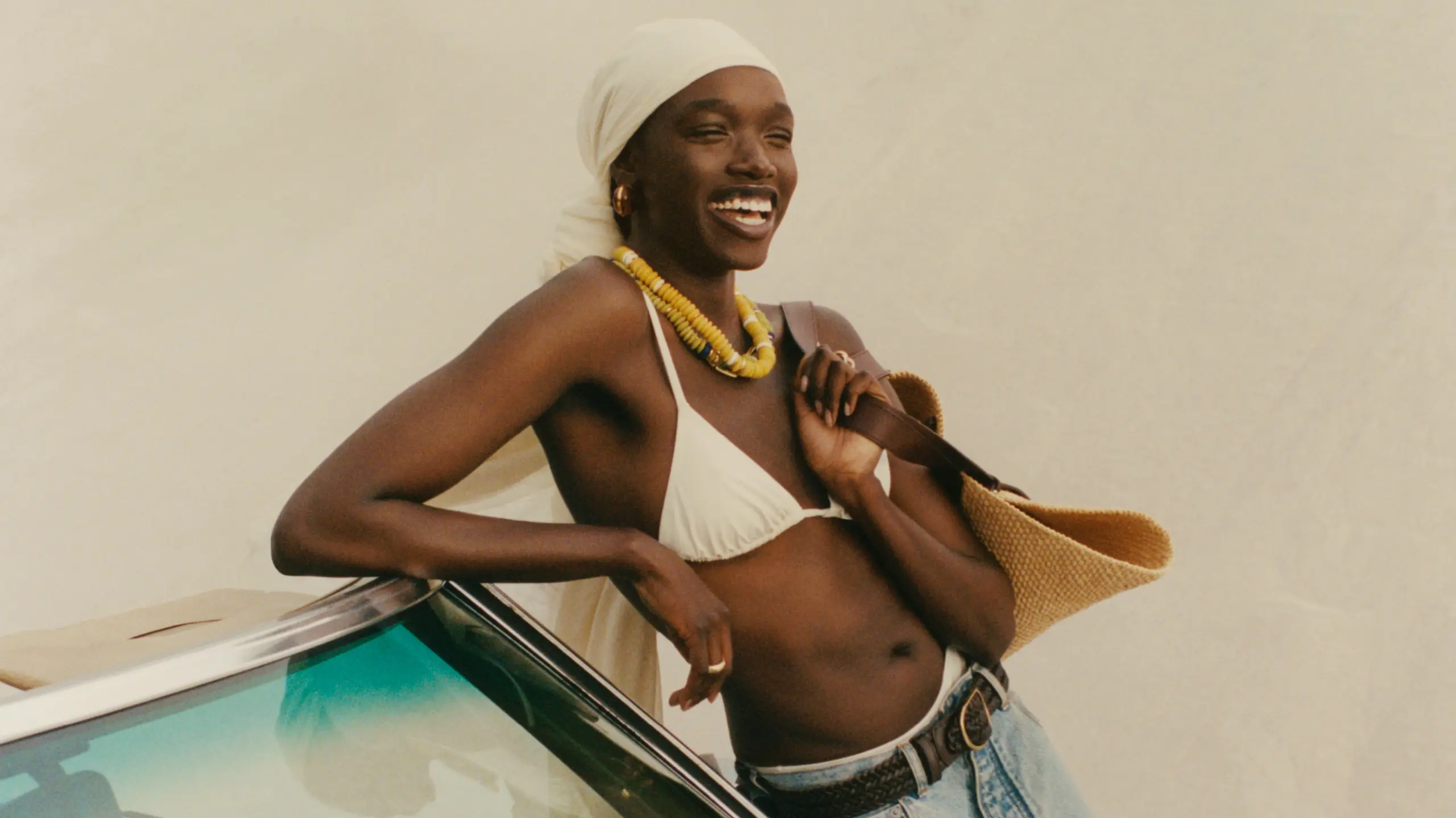 Eine Schwarze Frau in Jeansshorts, einem Bikinioberteil und einer Stofftasche, sie lacht und lehnt an einem Cabrio